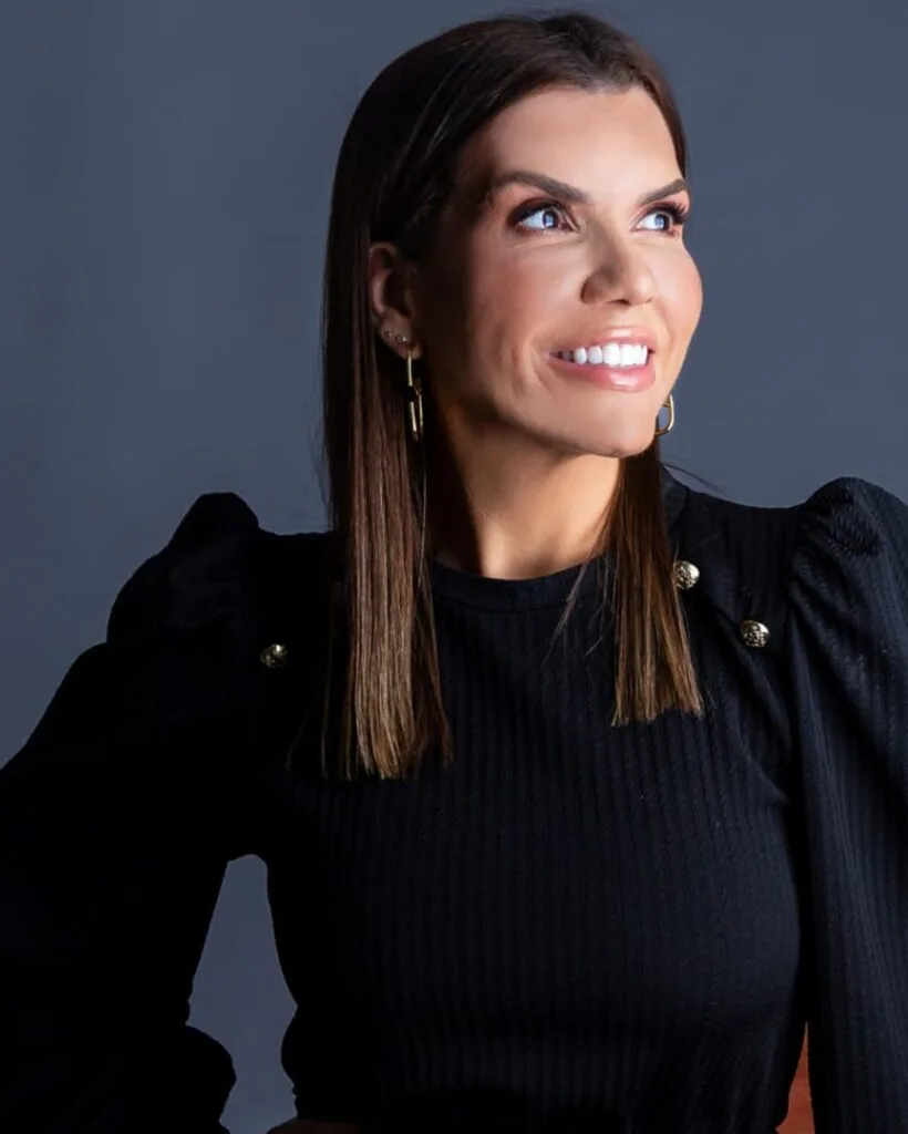 Dra. Gabriela Brum sorrindo, de blusa preta, diante de um fundo cinza-azulado
